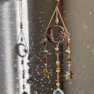 May include: A hanging suncatcher ornament featuring a gold-toned crescent moon and star charm, accented with amber, brown, and clear faceted beads. The design includes a dangling clear crystal sphere at the bottom, suspended by a gold-colored chain.