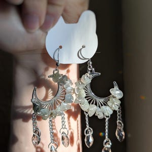 May include: A pair of silver-toned crescent moon earrings featuring small, tumbled, translucent grey-green stones set along the curve. Each earring includes three dangling silver chains ending in clear, faceted, teardrop-shaped crystal beads.