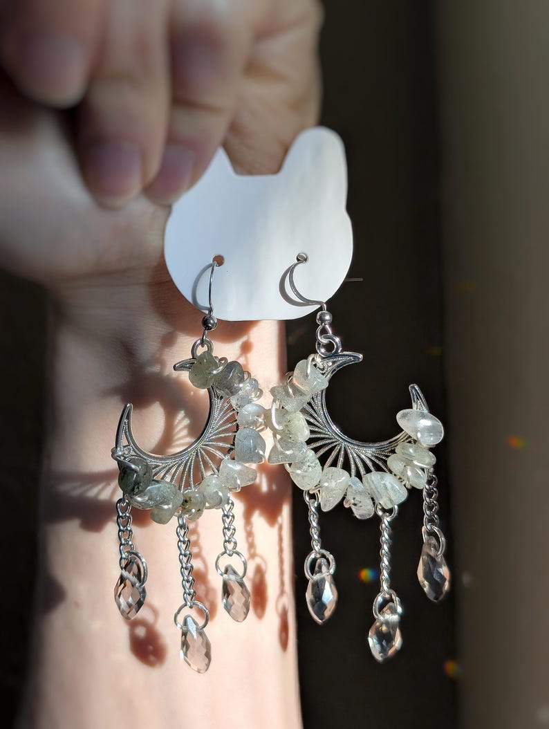 May include: A pair of silver-toned crescent moon earrings featuring small, tumbled, translucent grey-green stones set along the curve. Each earring includes three dangling silver chains ending in clear, faceted, teardrop-shaped crystal beads.