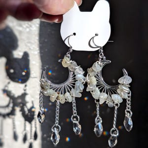 May include: A pair of silver-toned crescent moon earrings featuring wire-wrapped light-colored stone chips along the curve. Three small silver chains hang from each moon, ending in clear, faceted, teardrop-shaped crystal beads.