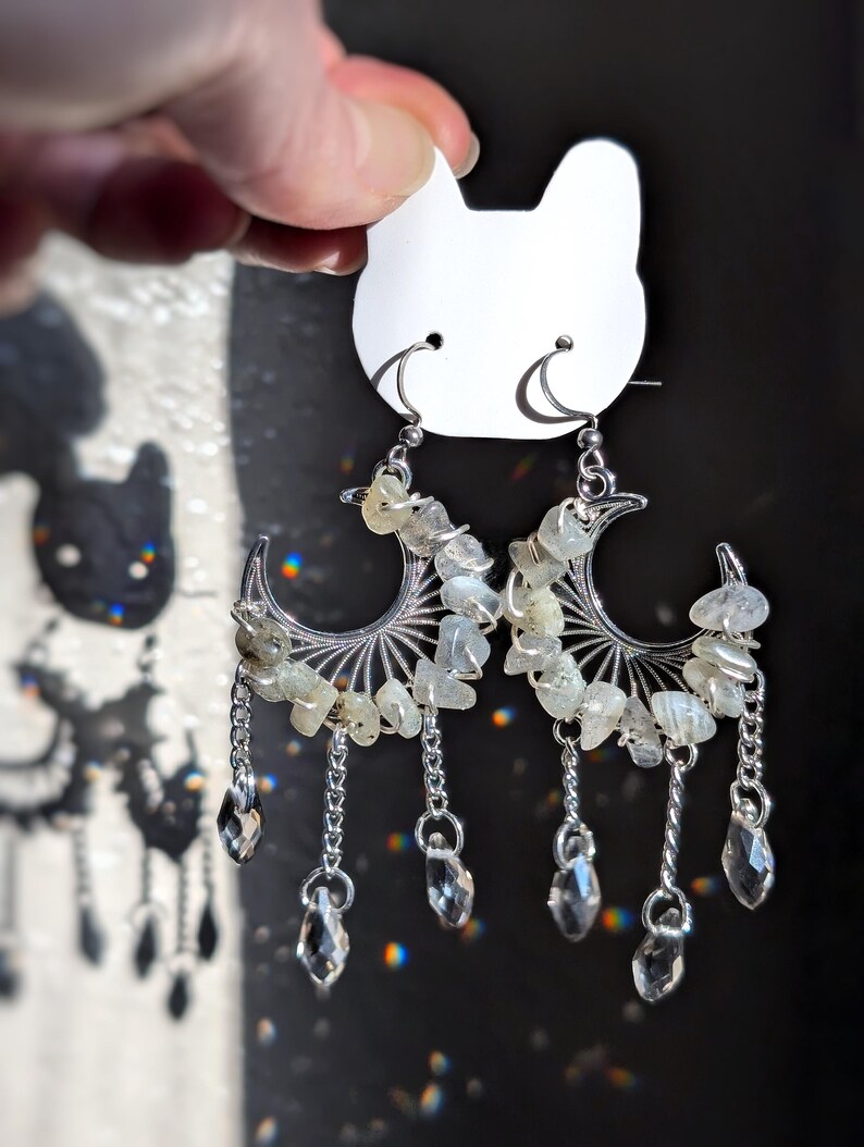 May include: A pair of silver-toned crescent moon earrings featuring wire-wrapped light-colored stone chips along the curve. Three small silver chains hang from each moon, ending in clear, faceted, teardrop-shaped crystal beads.