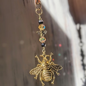 May include: A gold-colored bee charm keychain with a lobster clasp. The charm features a detailed bee pendant, iridescent beads, and a small gold-colored bee charm. The keychain is suspended from a gold-colored clasp.