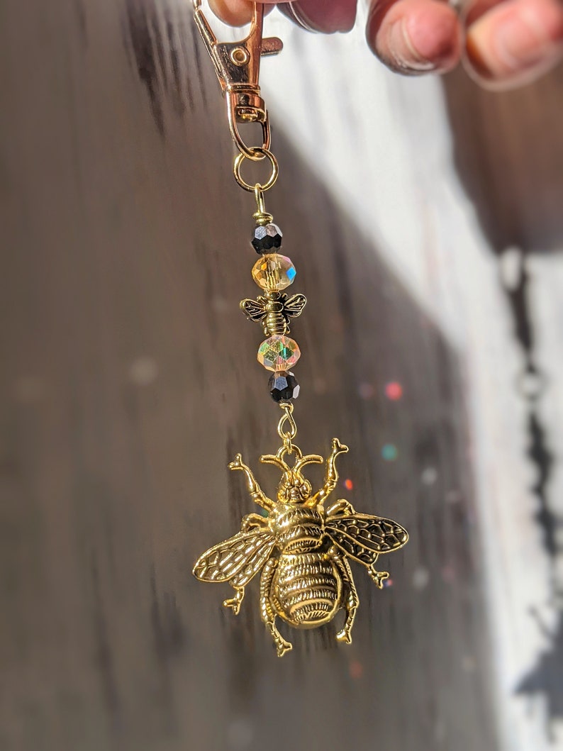 May include: A gold-colored bee charm keychain with a lobster clasp. The charm features a detailed bee pendant, iridescent beads, and a small gold-colored bee charm. The keychain is suspended from a gold-colored clasp.