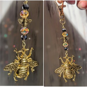 May include: A gold-colored bee charm keychain with a lobster clasp. The keychain features a large, detailed bee pendant, smaller bee charms, and iridescent and black beads. The design is suspended from a gold-tone chain.