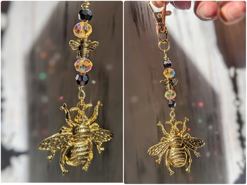 May include: A gold-colored bee charm keychain with a lobster clasp. The keychain features a large, detailed bee pendant, smaller bee charms, and iridescent and black beads. The design is suspended from a gold-tone chain.