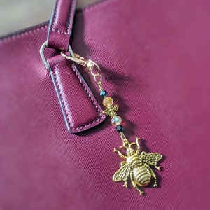 May include: A gold-colored bee charm with a beaded accent, attached to a burgundy handbag. The bee charm is detailed with intricate wings and body features. The beads are black, clear, and yellow. The handbag has a textured surface.