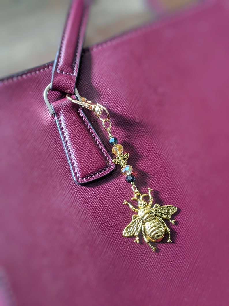 May include: A gold-colored bee charm with a beaded accent, attached to a burgundy handbag. The bee charm is detailed with intricate wings and body features. The beads are black, clear, and yellow. The handbag has a textured surface.