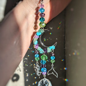 May include: A crystal suncatcher with a crescent moon design, featuring a variety of colorful beads, including turquoise, green, purple, and blue. The suncatcher also includes two silver mermaid tail charms and a large crystal ball.