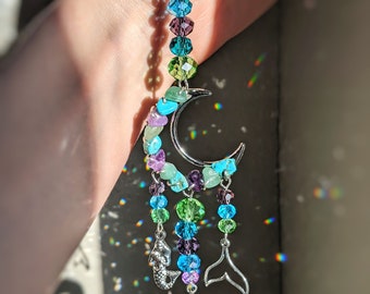 May include: A crystal suncatcher with a crescent moon design, featuring a variety of colourful beads, including turquoise, green, purple, and blue. The suncatcher also includes two silver mermaid tail charms and a large crystal ball.