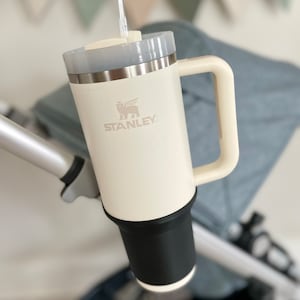 May include: A white Stanley travel mug with a black lid and a black handle. The mug has a logo that says "STANLEY" and a picture of a bird with wings spread. The mug is attached to a stroller.
