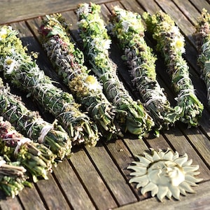 Könnte beinhalten: Neun Salbei-Bündel, jedes mit Bindfaden gebunden und mit getrockneten Blumen und Kräutern verziert, liegen auf einer verwitterten Holzoberfläche. Ein dekoratives sonnenförmiges Objekt ist ebenfalls vorhanden. Die Bündel sind etwa 15-20 cm lang.