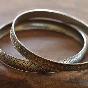 Könnte beinhalten: Zwei silberne Armreifen mit aufwendigen Designs. Der obere Armreif hat ein sich wiederholendes Kreis-Muster, während der untere Armreif ein geflochtenes Muster hat.