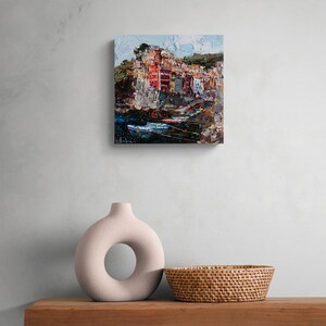 May include: An oil painting of a coastal village with vibrant buildings and water. The artwork uses a textured, impasto technique with visible brushstrokes. The painting is displayed on a wall above a wooden shelf with a decorative vase and a woven basket.