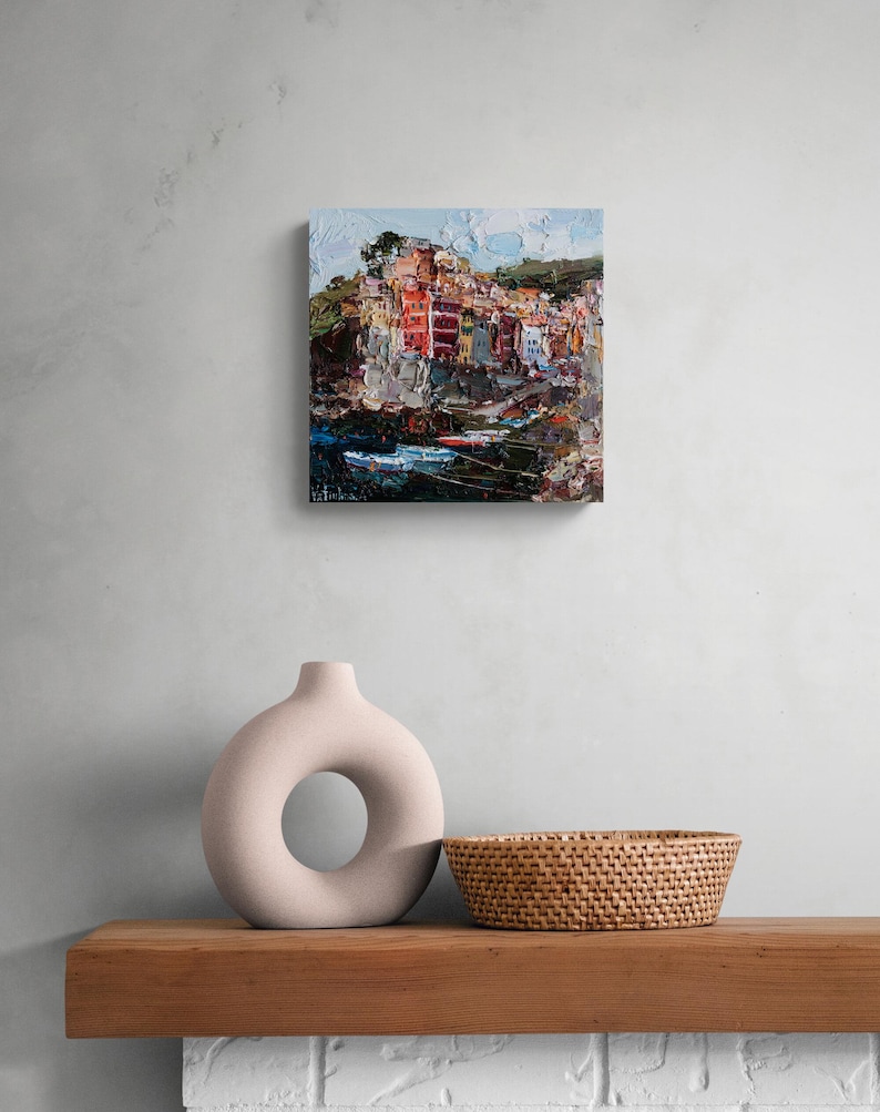 May include: An oil painting of a coastal village with vibrant buildings and water. The artwork uses a textured, impasto technique with visible brushstrokes. The painting is displayed on a wall above a wooden shelf with a decorative vase and a woven basket.