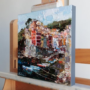 May include: An oil painting on canvas shows a vibrant coastal town with buildings in red, orange, and yellow. The artwork uses a textured, impasto technique, with visible brushstrokes. The painting is displayed on a wooden easel.