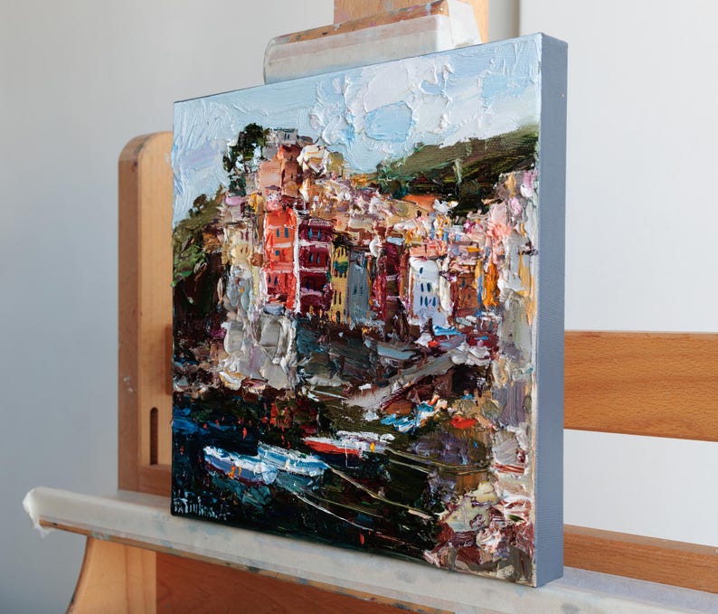 May include: An oil painting on canvas shows a vibrant coastal town with buildings in red, orange, and yellow. The artwork uses a textured, impasto technique, with visible brushstrokes. The painting is displayed on a wooden easel.