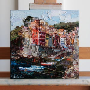 May include: Square oil painting depicting a coastal town with vibrant buildings and boats. The artwork uses an impasto technique with thick brushstrokes. The buildings are painted in shades of red, orange, yellow, and white, with a blue sky and green hills in the background.