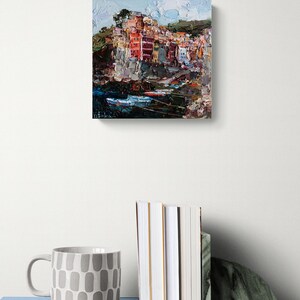 May include: An oil painting of a coastal village with vibrant buildings. The artwork uses an impressionistic style with textured brushstrokes and a palette of red, orange, and blue. The painting is on a white wall, with a mug and books in the foreground.