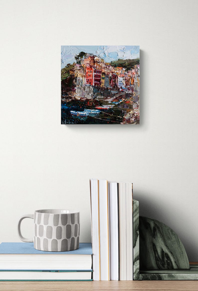 May include: An oil painting of a coastal village with vibrant buildings. The artwork uses an impressionistic style with textured brushstrokes and a palette of red, orange, and blue. The painting is on a white wall, with a mug and books in the foreground.