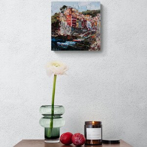 May include: An oil painting of a coastal village with vibrant buildings and boats. A white flower in a green glass vase, two plums, and a lit candle in a brown jar sit on a wooden table with a geometric design.