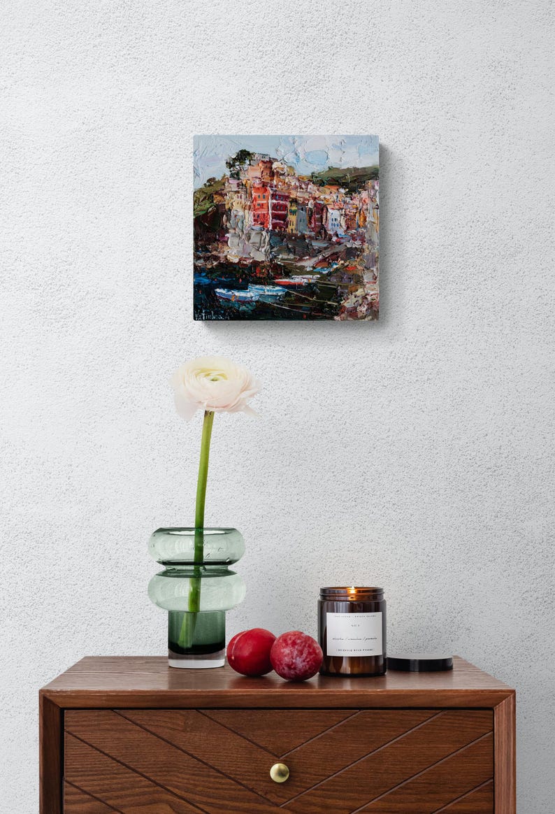 May include: An oil painting of a coastal village with vibrant buildings and boats. A white flower in a green glass vase, two plums, and a lit candle in a brown jar sit on a wooden table with a geometric design.