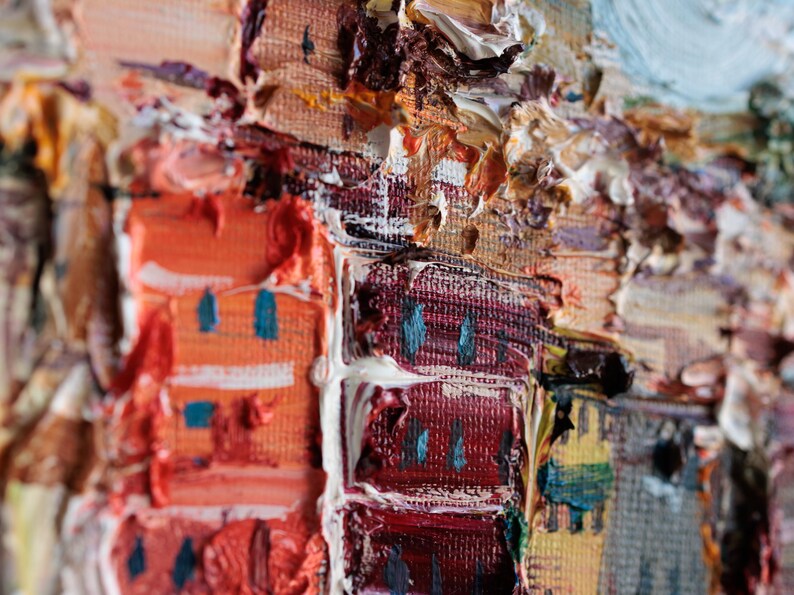 May include: Close-up of an oil painting showing a cityscape with textured brushstrokes. The artwork features buildings in shades of orange, red, and grey, with accents of blue and white. The painting style is impressionistic.
