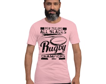 May include: A pale pink t-shirt featuring a black graphic. The design includes the text "NEW ZEALAND ALL BLACKS" above a rugby ball illustration and the word "Rugby" in a stylized font. Below the ball, the text reads "CHAMPIONS".
