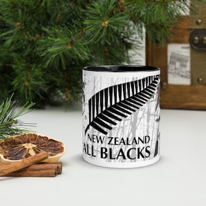 以下が含まれることがあります： 黒い内側の白いセラミックマグ。黒いシダの葉のデザインと「NEW ZEALAND ALL BLACKS」の文字が特徴です。マグカップは黒い縁と、灰色の木のような模様の白い外観をしています。
