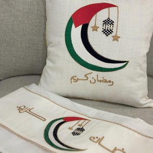 May include: White cushion and runner set featuring an embroidered crescent moon design in red, green, black, and white. The design includes hanging lanterns and stars, with gold Arabic text. The cushion has gold trim.