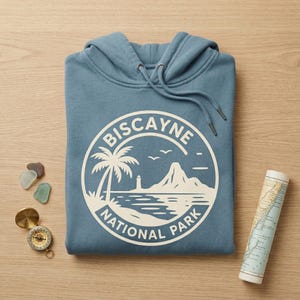 May include: A blue hoodie with a white graphic of Biscayne National Park. The design includes a palm tree, lighthouse, mountain, and the text "Biscayne National Park". A compass, map, and sea glass are next to the hoodie.
