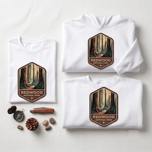May include: A white t-shirt, a white hoodie, and a white sweatshirt, each featuring a hexagonal patch with a forest illustration and the text Redwood National Park. Also included are a compass, a pocket knife, a pinecone, and small stones.