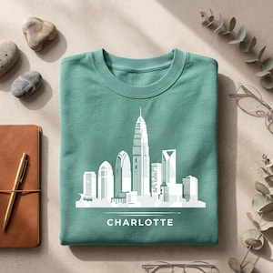 May include: A teal crewneck sweatshirt featuring a white graphic print of the Charlotte city skyline. The design includes a stylized illustration of various skyscrapers and the word CHARLOTTE printed in white capital letters below the skyline.