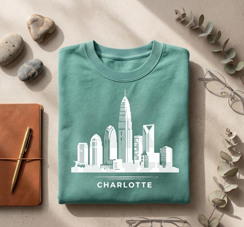 May include: A teal crewneck sweatshirt featuring a white graphic print of the Charlotte city skyline. The design includes a stylized illustration of various skyscrapers and the word CHARLOTTE printed in white capital letters below the skyline.