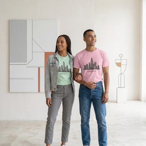 May include: A light green t-shirt and a pink t-shirt, both featuring a dark gray graphic of the Charlotte skyline and the text "CHARLOTTE" printed underneath. The shirts are worn with a light blue denim jacket, gray trousers, and blue jeans.