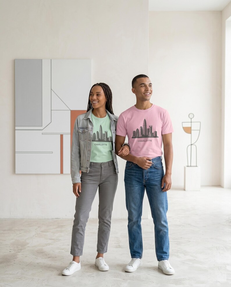 May include: A light green t-shirt and a pink t-shirt, both featuring a dark gray graphic of the Charlotte skyline and the text "CHARLOTTE" printed underneath. The shirts are worn with a light blue denim jacket, gray trousers, and blue jeans.