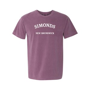 May include: A mauve-colored t-shirt with the word "SIMONDS" arched across the chest in white capital letters. Below it, the text "NEW BRUNSWICK" is also in white. The shirt has short sleeves and a crew neck.