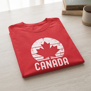 May include: A bright red t-shirt featuring a white distressed circular graphic with a maple leaf in the center and the word CANADA printed in bold white capital letters below the circle. The shirt is folded neatly on a light-colored wooden surface.