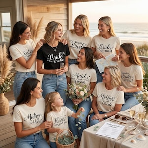 May include: A group of women and a young girl wearing matching t-shirts with wedding-related text. The shirts are cream-coloured and black, with phrases like "Bride to be," "Maid of Honour," and "Flower Girl." They are gathered outdoors near the beach.