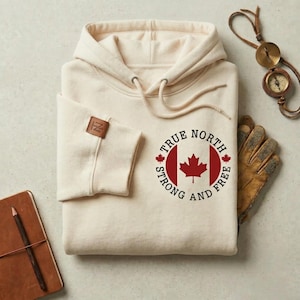 May include: A cream-coloured hoodie with a drawstring hood and a circular graphic featuring a red maple leaf and the words "True North Strong and Free." A leather patch with the initials "IZ" is on the sleeve. A compass, gloves, and a notebook are also visible.