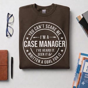 May include: A brown sweatshirt with a white circular graphic that reads "YOU CAN'T SCARE ME - I'M A CASE MANAGER - I'VE HEARD IT, SEEN IT & WRITTEN A GOAL FOR IT". The sweatshirt is folded on a light surface.