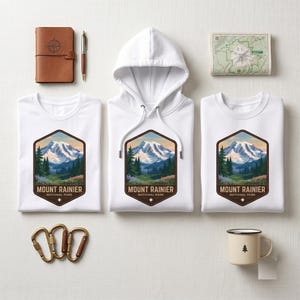 May include: Two white t-shirts and one white hooded sweatshirt featuring a graphic of Mount Rainier National Park, a brown leather journal, a pen, a map, three metal carabiners, and a cream-colored enamel mug with a small tree icon.