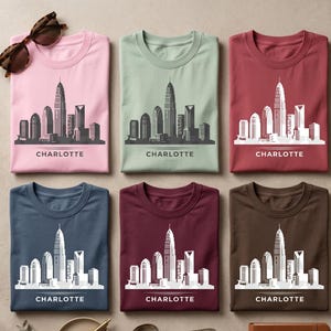 May include: Six folded t-shirts in pink, sage green, maroon, navy blue, dark red, and brown, each featuring a white graphic of the Charlotte skyline and the text "CHARLOTTE" printed below. A pair of brown tortoiseshell sunglasses sits at the top left.