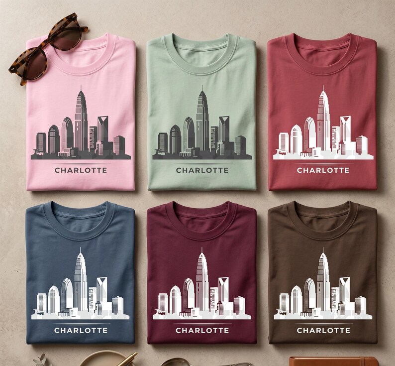 May include: Six folded t-shirts in pink, sage green, maroon, navy blue, dark red, and brown, each featuring a white graphic of the Charlotte skyline and the text "CHARLOTTE" printed below. A pair of brown tortoiseshell sunglasses sits at the top left.