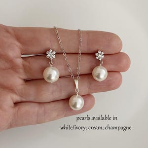 May include: A pearl jewelry set featuring a necklace and matching earrings. The necklace has a single pearl pendant, and the earrings have pearl drops. The earrings are topped with a flower-shaped design. The text says "pearls available in white/ivory; cream; champagne."