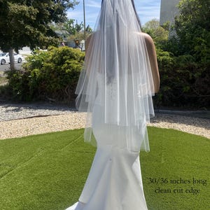 May include: A two-tier white bridal veil with a clean cut edge, measuring 30/36 inches long, worn over a white floor-length mermaid-style wedding dress with a corset-style back closure and a long train extending across the ground.