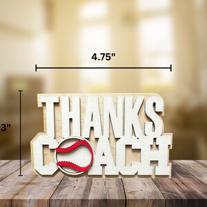 Puede incluir: Se&ntilde;al de madera con las palabras "Thanks Coach" en letras blancas. Una pelota de b&eacute;isbol roja y blanca est&aacute; en el medio de la se&ntilde;al. La se&ntilde;al mide 12,1 cm de ancho y 7,6 cm de alto.