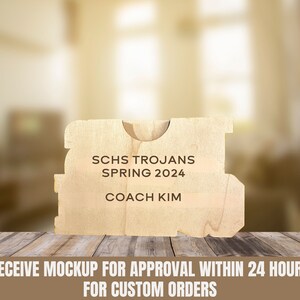 May include: A light-colored wooden plaque with the text "SCHS TROJANS SPRING 2024 COACH KIM" in black. The plaque has a unique shape with a curved top and cut-out details. The plaque is on a wooden surface.