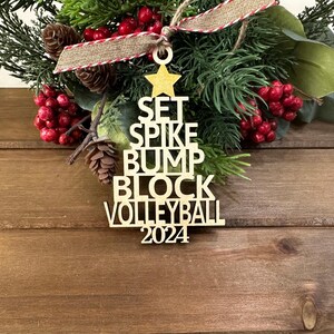 Könnte beinhalten: Ein hölzerner Weihnachtsbaum-Anhänger mit den Worten "SET SPIKE BUMP BLOCK VOLLEYBALL 2024" darauf. Der Anhänger ist golden und hat einen Stern oben drauf.