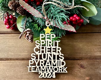 Puede incluir: Adorno de árbol de Navidad de madera con las palabras "PEP SPIRIT CHEER STUNTS PYRAMID TEAMWORK 2024" en letras negras. El adorno está decorado con una estrella dorada.