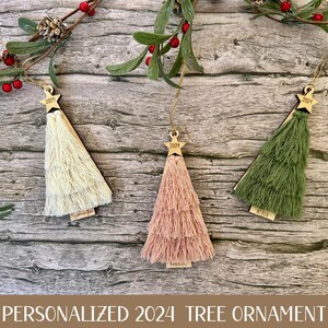 May include: Three personalized Christmas tree ornaments with a tassel fringe. The ornaments are made of wood and are painted in white, pink, and green. Each ornament has a star on top and the year 2024 written on it.